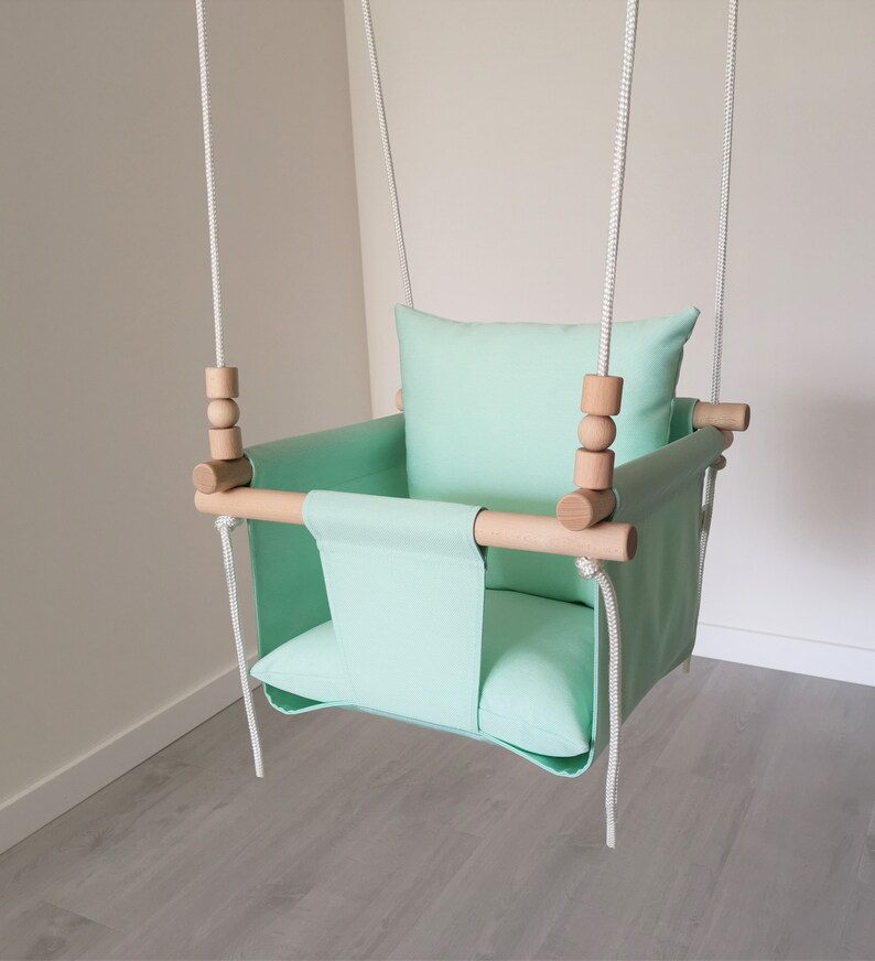 May include: A mint green baby swing with a wooden frame and white ropes. The swing has a padded seat and a pillow.