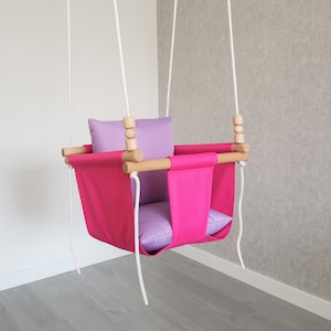 Puede incluir: Un asiento de columpio de tela rosa con un cojín morado, colgado de una cuerda blanca con cuentas de madera.