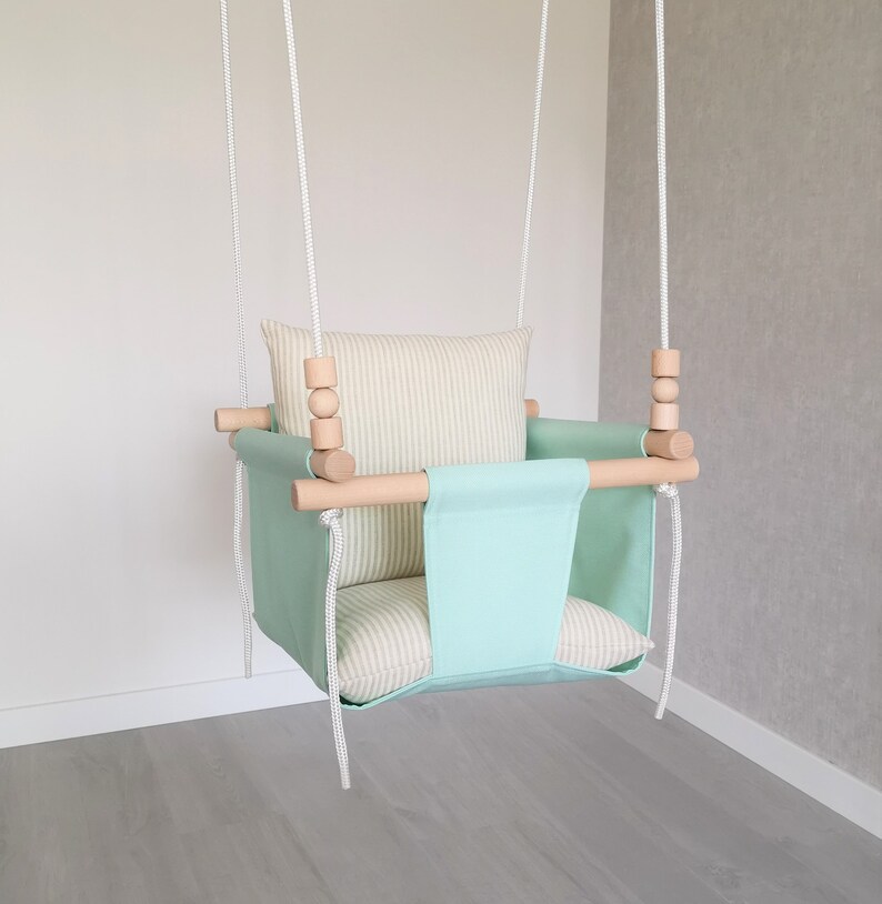 May include: A light green baby swing with a striped cushion and wooden bars. The swing is hanging from white ropes.