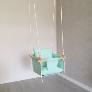 May include: A light green baby swing with a wooden frame and white ropes. The swing has a padded seat and a pillow.