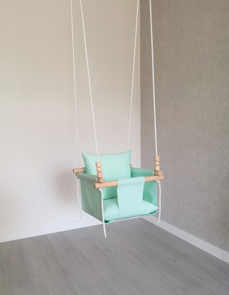 May include: A light green baby swing with a wooden frame and white ropes. The swing has a padded seat and a pillow.