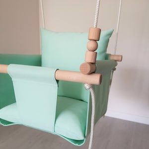 May include: A light green fabric baby swing with a wooden frame and wooden beads. The swing has a padded seat and back.