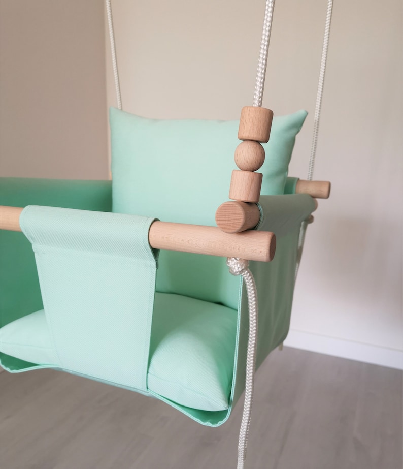May include: A light green fabric baby swing with a wooden frame and wooden beads. The swing has a padded seat and back.