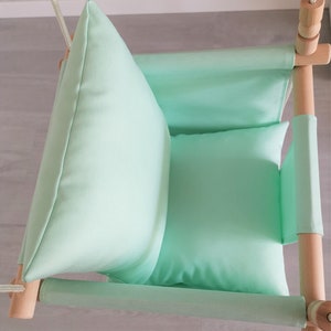 May include: A light green fabric baby swing with wooden frame and rope. The swing has two cushions for added comfort.