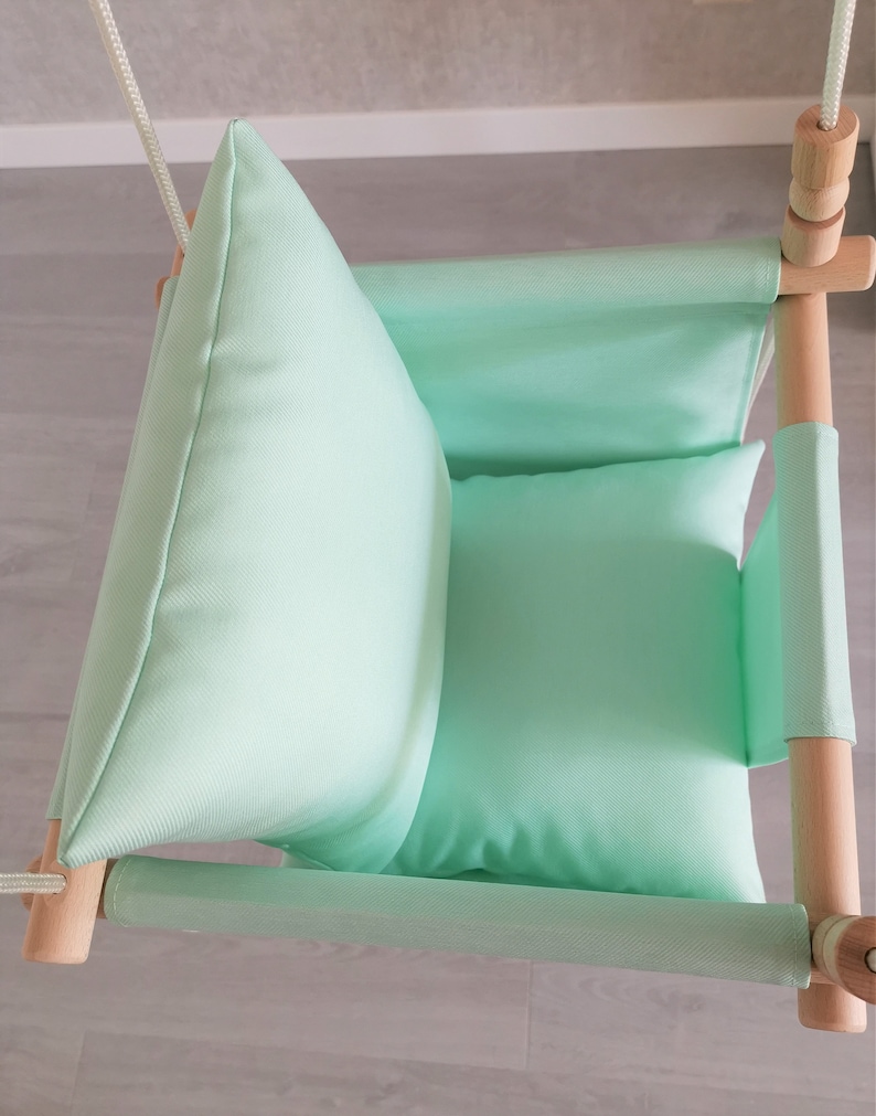 May include: A light green fabric baby swing with wooden frame and rope. The swing has two cushions for added comfort.