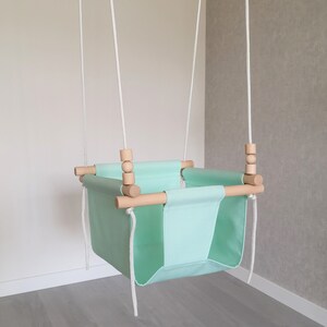 May include: A light green baby swing with wooden bars and white ropes. The swing is hanging from the ceiling.