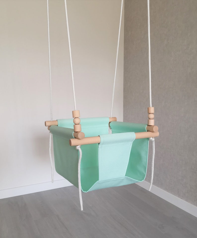 May include: A light green baby swing with wooden bars and white ropes. The swing is hanging from the ceiling.