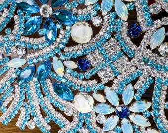 May include: Close-up of a decorative object with a floral design. The piece features a combination of blue, turquoise, and clear crystals and gemstones. The design includes large, faceted blue gems and smaller, clear crystals.