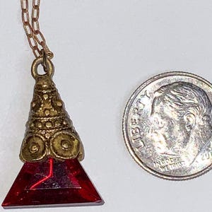 May include: A vintage pendant necklace featuring a red, triangular gemstone. The pendant has a decorative, gold-toned metal cap and hangs from a delicate chain. The necklace is shown next to a U.S. dime for size comparison.