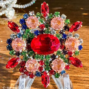 May include: A vintage brooch with a floral design. The brooch features a large, oval red gemstone at the center, surrounded by pink, green, blue, and red gemstones. The brooch is set in a gold-tone metal and has a clear plastic support.