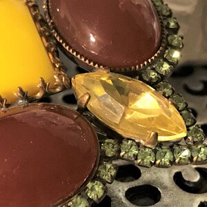 May include: Close-up of a vintage brooch with a floral design. The brooch features large, oval and rectangular stones in shades of yellow and brown, accented with smaller green rhinestones and gold-tone metal detailing.