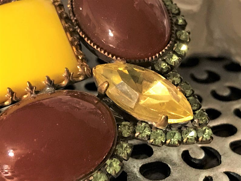 May include: Close-up of a vintage brooch with a floral design. The brooch features large, oval and rectangular stones in shades of yellow and brown, accented with smaller green rhinestones and gold-tone metal detailing.
