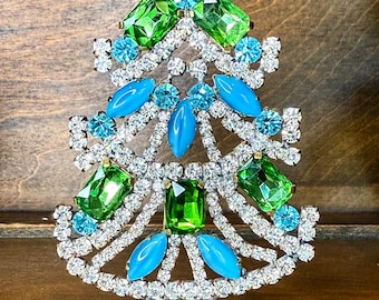 May include: A decorative Christmas tree figurine with a metal base. The tree is adorned with clear, green, and turquoise-colored gemstones. The base is a gold-colored metal with an ornate design. The tree is approximately 5 inches tall.