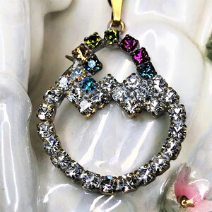 May include: A gold pendant with a heart-shaped design, featuring a circle of clear rhinestones and a smaller heart-shaped design with colorful rhinestones.