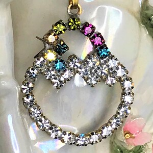 May include: A gold pendant with a heart-shaped design. The heart is made of clear rhinestones and has a smaller circle inside with colorful rhinestones.