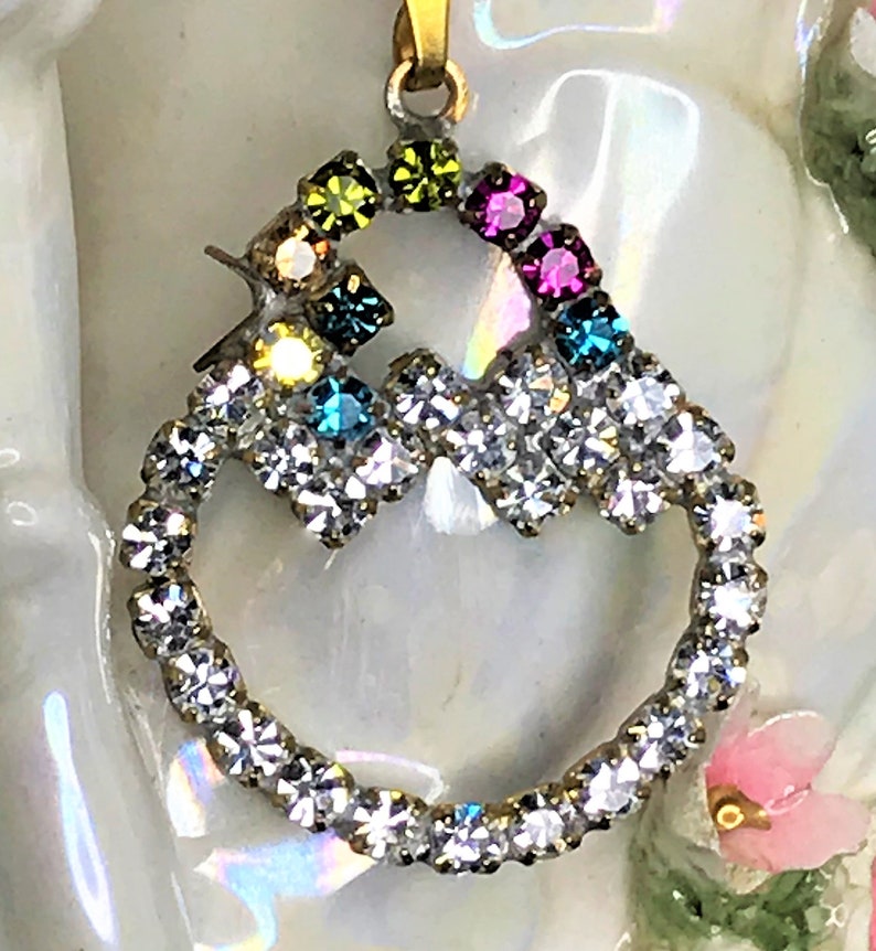 May include: A gold pendant with a heart-shaped design. The heart is made of clear rhinestones and has a smaller circle inside with colorful rhinestones.