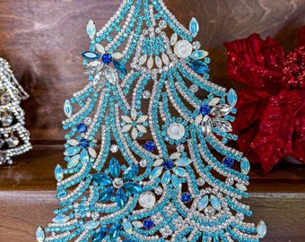 May include: A decorative Christmas tree sculpture with a turquoise and silver design. The tree is adorned with various blue and white gemstones and sits on a gold-colored base. The tree is a festive holiday decoration.