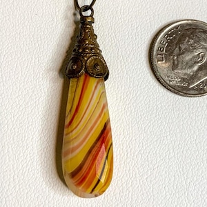 May include: A teardrop-shaped pendant with a swirling pattern of yellow, orange, and red. The pendant is topped with a decorative bronze cap and hangs from a thin chain. A dime is next to the pendant for size comparison.