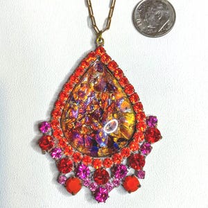 May include: A teardrop-shaped pendant necklace with a gold chain. The pendant features a mosaic of orange, purple, and gold colors, framed by red and pink rhinestones. The necklace is a vintage costume jewelry piece.