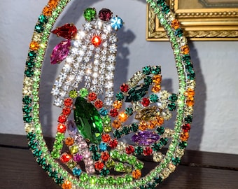 May include: A colorful oval-shaped ornament made of rhinestones and glass beads. The ornament features a variety of colors, including green, red, orange, yellow, purple, and blue. The ornament is surrounded by a silver-colored metal frame.