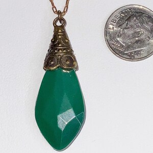May include: A vintage-style pendant necklace featuring a faceted, teardrop-shaped, emerald green gemstone. The pendant is suspended from a delicate gold-colored chain and has a decorative, ornate cap. A U.S. dime is next to the pendant for size comparison.