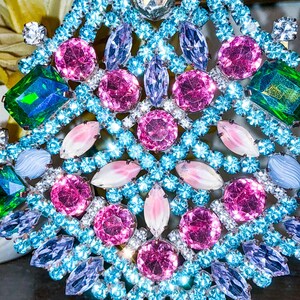 May include: Close-up of a sparkling, ornate brooch with a symmetrical design. The brooch features a variety of colorful gemstones, including pink, blue, green, and clear crystals. The gemstones are set in a light blue metal frame, creating a dazzling effect.