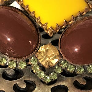 May include: Close-up of a vintage brooch featuring large, oval, brown cabochons and a yellow gemstone. The brooch is accented with small, light green rhinestones and gold-toned metal detailing. The background is a silver, open-work metal.