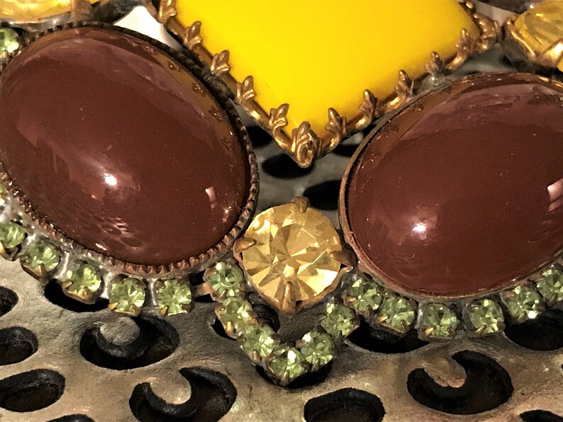 May include: Close-up of a vintage brooch featuring large, oval, brown cabochons and a yellow gemstone. The brooch is accented with small, light green rhinestones and gold-toned metal detailing. The background is a silver, open-work metal.