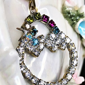 May include: A gold pendant with a heart-shaped design, featuring a ring of clear rhinestones and a smaller heart-shaped design with colorful rhinestones in shades of green, blue, pink, and purple.