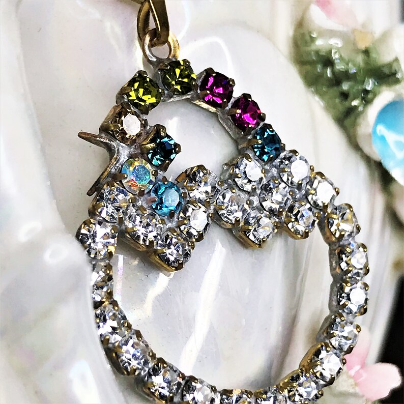 May include: A gold pendant with a heart-shaped design, featuring a ring of clear rhinestones and a smaller heart-shaped design with colorful rhinestones in shades of green, blue, pink, and purple.