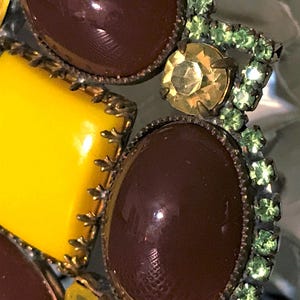 May include: Close-up of a vintage brooch with a mix of colors and textures. The brooch features large, glossy, oval and square-shaped brown and yellow stones, accented with smaller green and yellow rhinestones. The metalwork has an antique bronze finish.