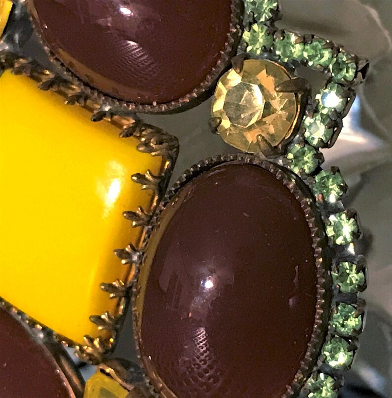May include: Close-up of a vintage brooch with a mix of colors and textures. The brooch features large, glossy, oval and square-shaped brown and yellow stones, accented with smaller green and yellow rhinestones. The metalwork has an antique bronze finish.