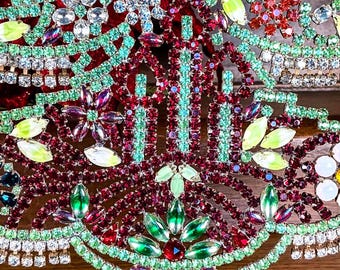 May include: Close-up of a decorative, ornate piece with a festive design. The piece features a combination of red, green, and clear rhinestones arranged in a floral and geometric pattern. The design is intricate and detailed, with a vintage aesthetic.
