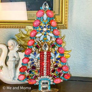 May include: A decorative Christmas tree figurine adorned with pink, blue, and clear gemstones. The tree features a base of colorful gems and a central column of red and white stones. A gold-framed picture is in the background.