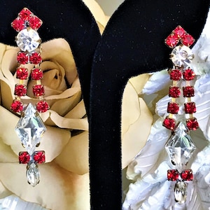 May include: A pair of dangle earrings with red and clear rhinestones. The earrings are on a black velvet display stand.