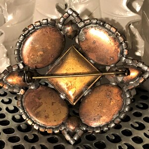 May include: Vintage brooch with a geometric design. The brooch features four oval-shaped copper-toned stones, a central square gold-toned stone, and a border of clear rhinestones. The pin backing is gold-toned.
