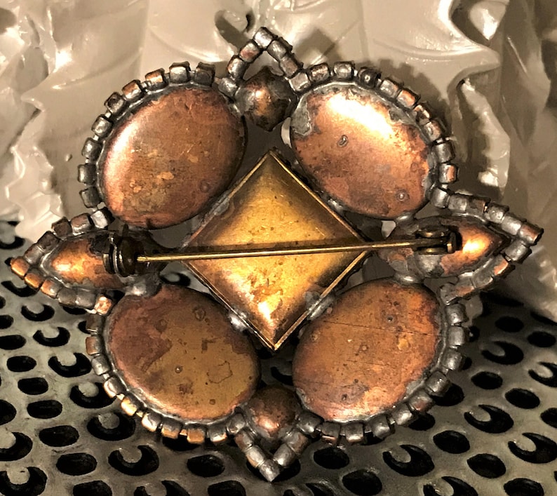 May include: Vintage brooch with a geometric design. The brooch features four oval-shaped copper-toned stones, a central square gold-toned stone, and a border of clear rhinestones. The pin backing is gold-toned.