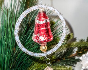 May include: A red glass bell ornament with white accents hangs from a silver chain and a clear plastic ring. The ornament is surrounded by green pine branches.