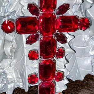 May include: A cross-shaped brooch featuring numerous faceted red glass stones in various shapes, including rectangular, round, and marquise cuts, set in a metal frame. The text "© Me and Moma" is visible in the bottom left corner of the image.