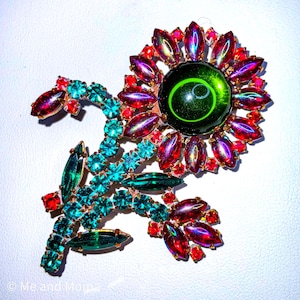 May include: A vintage floral brooch with a large green center stone and surrounding red and purple stones. The stem and leaves are adorned with turquoise and green stones. The brooch is set in a gold-tone metal.