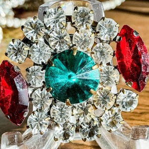 May include: A vintage-style brooch with a central teal-colored gemstone surrounded by clear, sparkling rhinestones. Two red, marquise-cut stones flank the central design. The brooch is set on a gold-toned metal base, creating a dazzling, eye-catching accessory.
