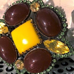 May include: A vintage brooch with a central yellow square surrounded by four oval brown stones. The brooch is accented with yellow and green rhinestones, set in a metal frame. The design is ornate and detailed.