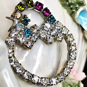 May include: A gold-toned ring with a circle of clear rhinestones and a smaller cluster of clear rhinestones. The ring is decorated with a row of colorful rhinestones in shades of green, blue, pink, and purple.