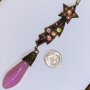 May include: A vintage-style pendant necklace featuring a shooting star design. The pendant is adorned with iridescent and purple rhinestones, and a teardrop-shaped pink stone. The metal has a bronze finish. A dime is included for size reference.