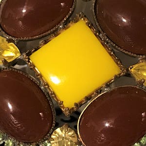 May include: Close-up of a vintage brooch with a central yellow square stone surrounded by brown oval stones. The brooch has a gold-tone metal frame and is accented with yellow and green rhinestones, creating a striking, ornate design.
