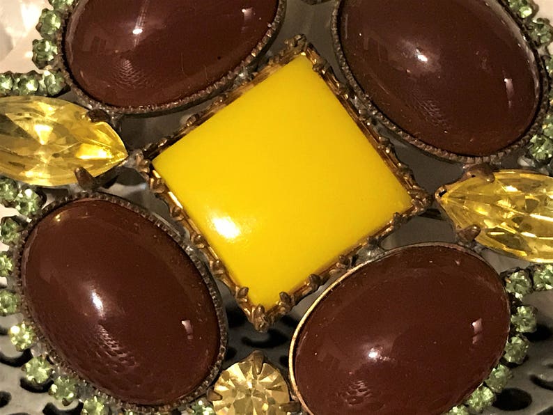 May include: Close-up of a vintage brooch with a central yellow square stone surrounded by brown oval stones. The brooch has a gold-tone metal frame and is accented with yellow and green rhinestones, creating a striking, ornate design.