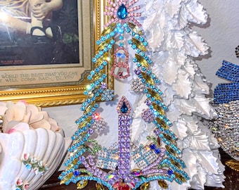 May include: A decorative Christmas tree figurine made of colorful rhinestones. The tree features blue, green, pink, and purple stones, with a gold base and a star on top. The tree is adorned with a candle and gift box accents. The text "Me and Moma" is visible.