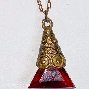 May include: A gold-colored pendant necklace with a red triangular gemstone. The pendant has an ornate, textured design and is suspended from a delicate chain. The gemstone is a deep red color and is cut into a triangular shape.