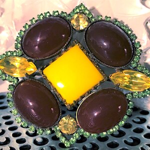 May include: A vintage brooch with a starburst design. It features a central yellow square surrounded by four oval, dark brown stones and four yellow, leaf-shaped stones. The brooch is trimmed with green rhinestones.