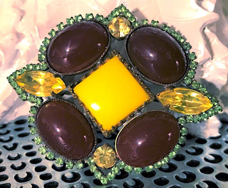 May include: A vintage brooch with a starburst design. It features a central yellow square surrounded by four oval, dark brown stones and four yellow, leaf-shaped stones. The brooch is trimmed with green rhinestones.